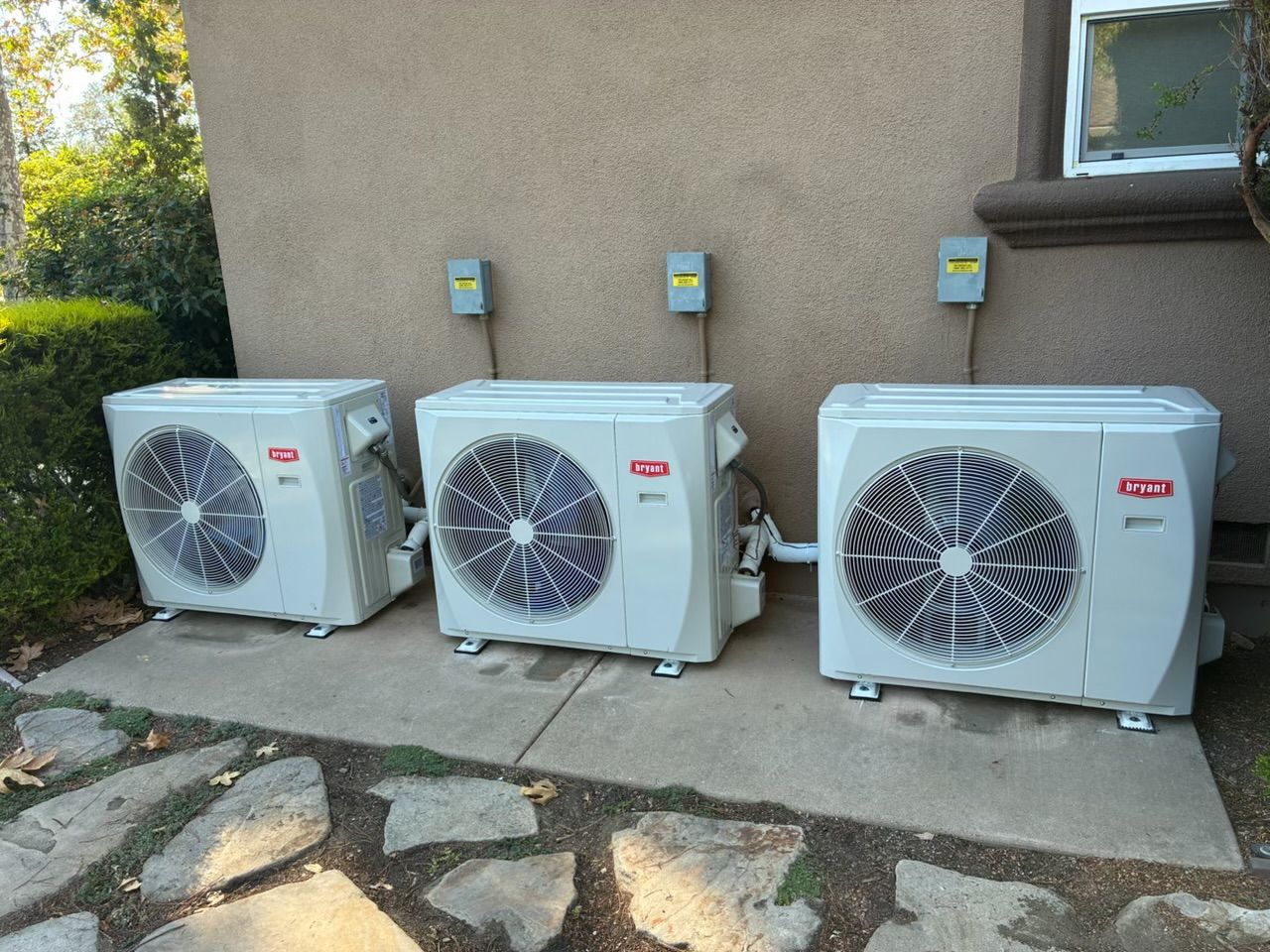 Three outdoor HVAC units beside a house wall, with meters above and stone patio in front.
