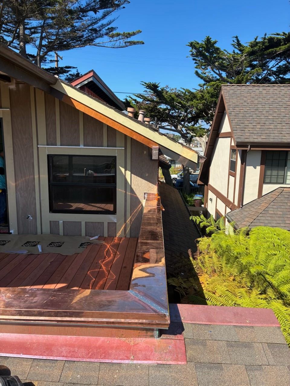 Narrow rooftop walkway between two houses, with wooden deck and shingled roofs under a blue sky.