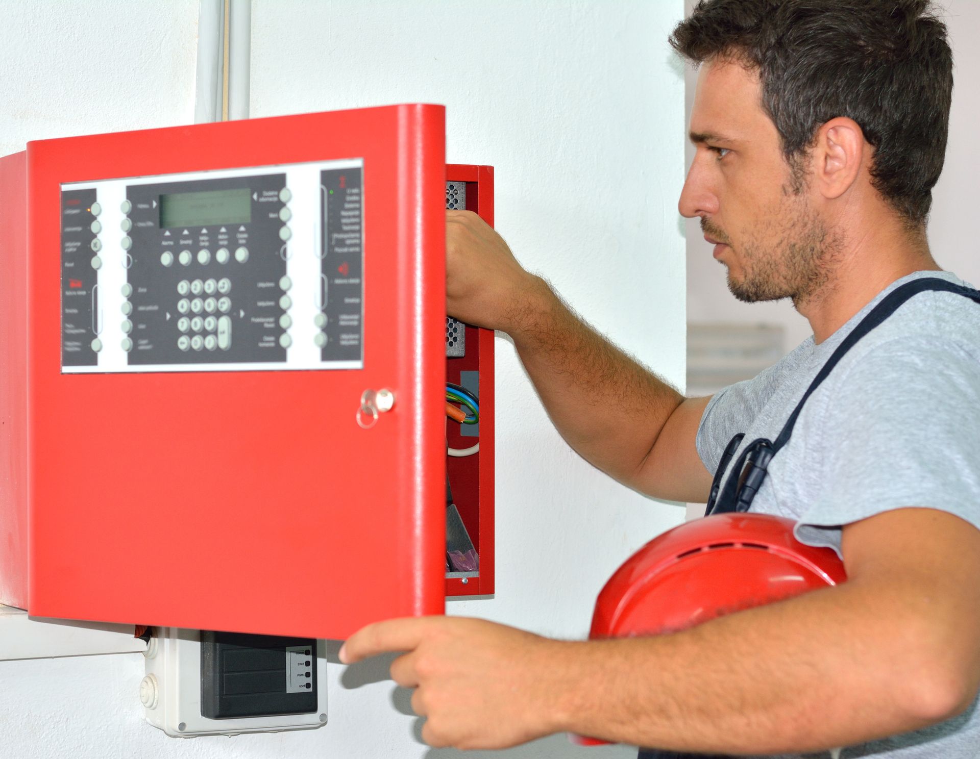 Man in overalls, inspecting a red fire alarm control panel. Holds a red hard hat, indoors.