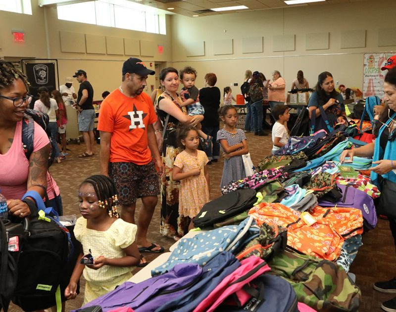 People at a back-to-school event selecting backpacks from a table, inside a community center.