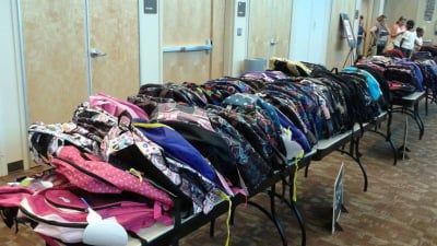 Rows of backpacks on tables indoors, likely for school supplies distribution.