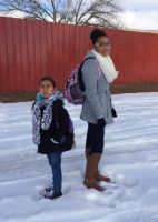 Two people stand in snow near a red wall. They wear jackets, backpacks, and boots.