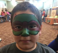 Boy with Teenage Mutant Ninja Turtle face paint, in a room with tables and people.