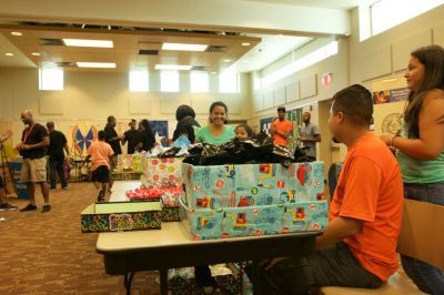 People at a gift distribution event inside a brightly lit room. Boxes of wrapped presents are on tables.