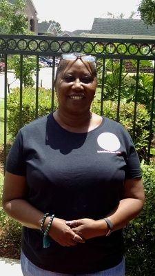 Woman standing in front of a black fence. She wears a black shirt, has sunglasses on her head, and smiles.