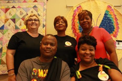 Group of five people posing in front of colorful quilts.