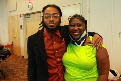 Man with dreadlocks in a blazer and a woman in a green top smile, arm-in-arm in a brightly lit room.
