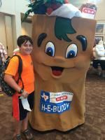 Boy in orange shirt with H-E-Buddy mascot. Both smile in front of store interior.