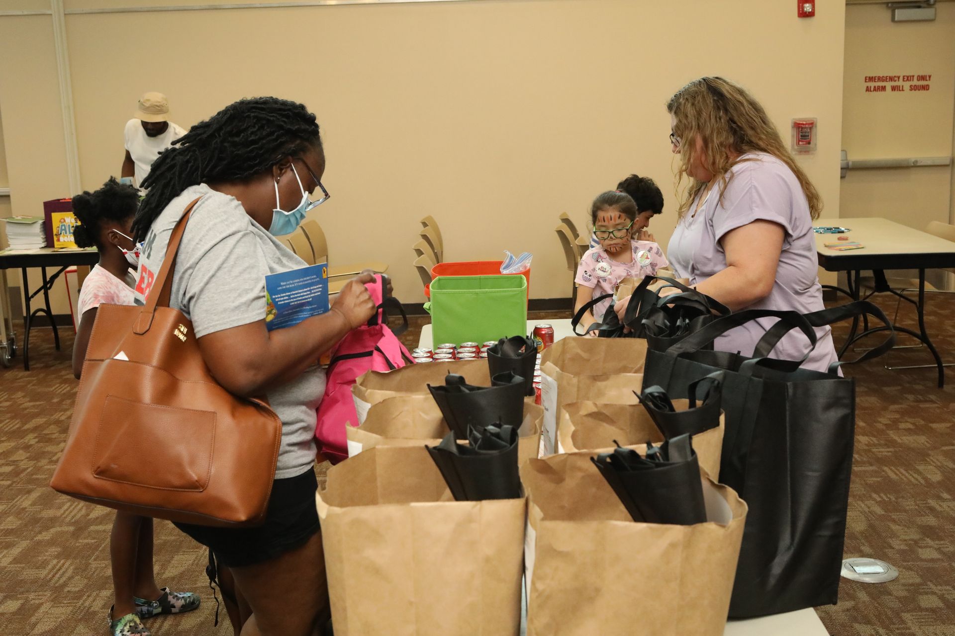 People packing bags with supplies at a table. Children watch. Neutral interior setting.
