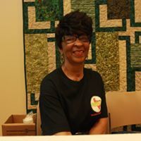 Woman in black shirt smiles, wearing glasses, in front of a green and brown quilt.