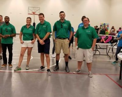 Group of people in green shirts, posing in a gym with a basketball hoop.