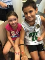 Two smiling children sit with temporary tattoos, a girl in pink, a boy in a basketball jersey.