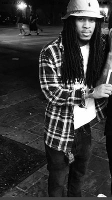 Person with dreadlocks, wearing a plaid shirt, bucket hat, and jeans, standing outside at night.