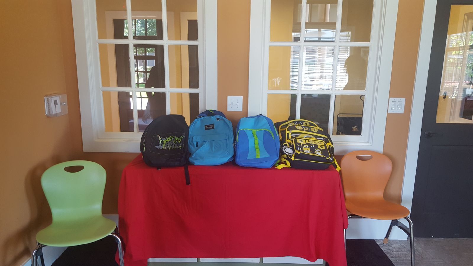 Four backpacks on a red table in front of windows, flanked by a green and orange chair.