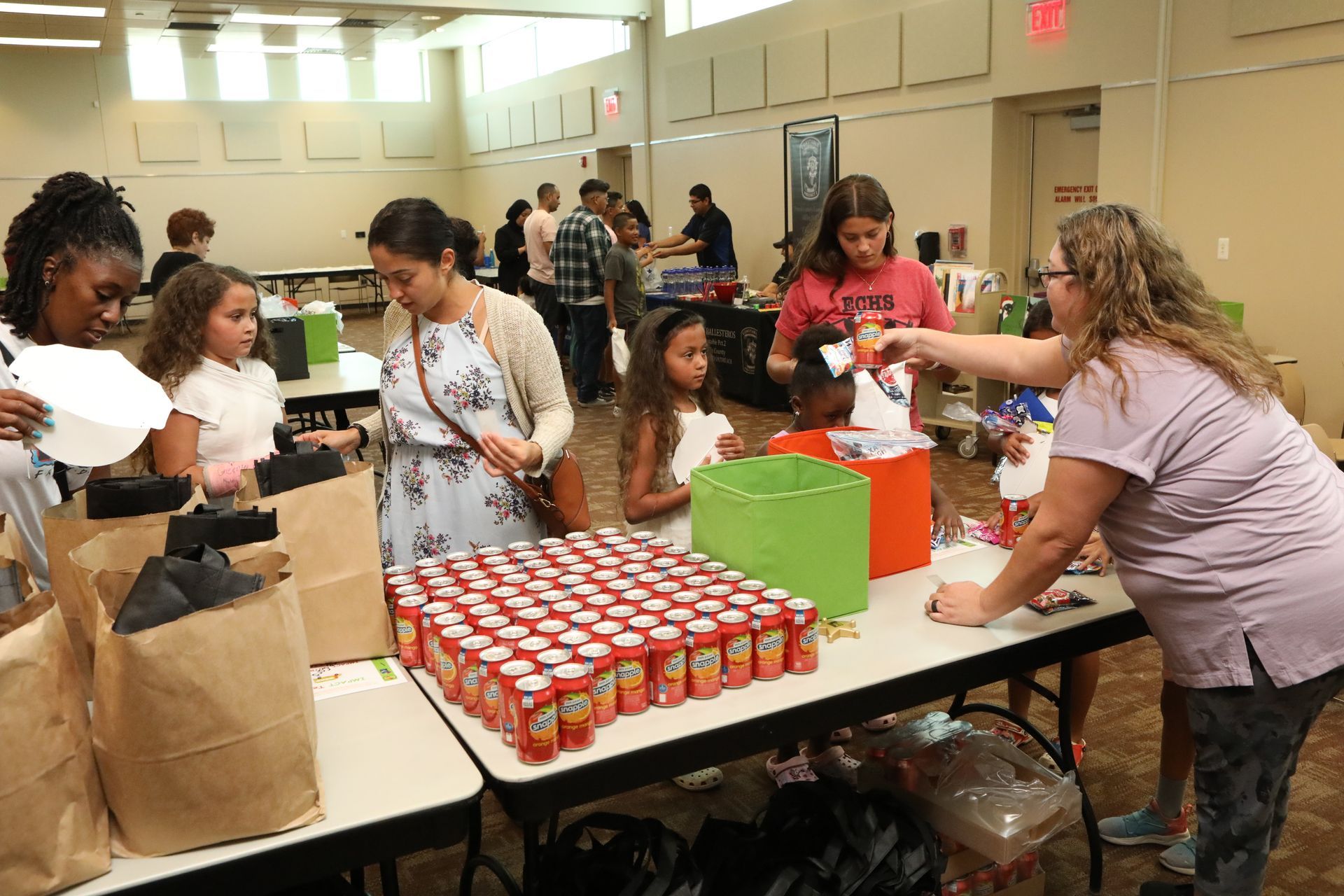People at tables filling bags with food and drinks in a brightly lit room.
