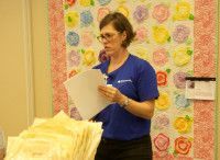 Woman in blue shirt holding papers, standing in front of a colorful floral quilt.