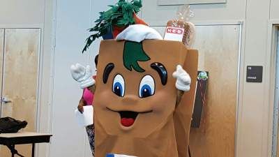 H-E-B grocery bag mascot smiles and waves, holding a paper; produce and bread displayed.