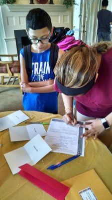 Boy in glasses watches woman sign paperwork at a table with cards.
