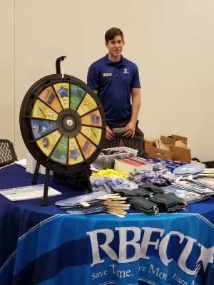 Man at a table with a prize wheel, RBFCU logo, and promotional materials.