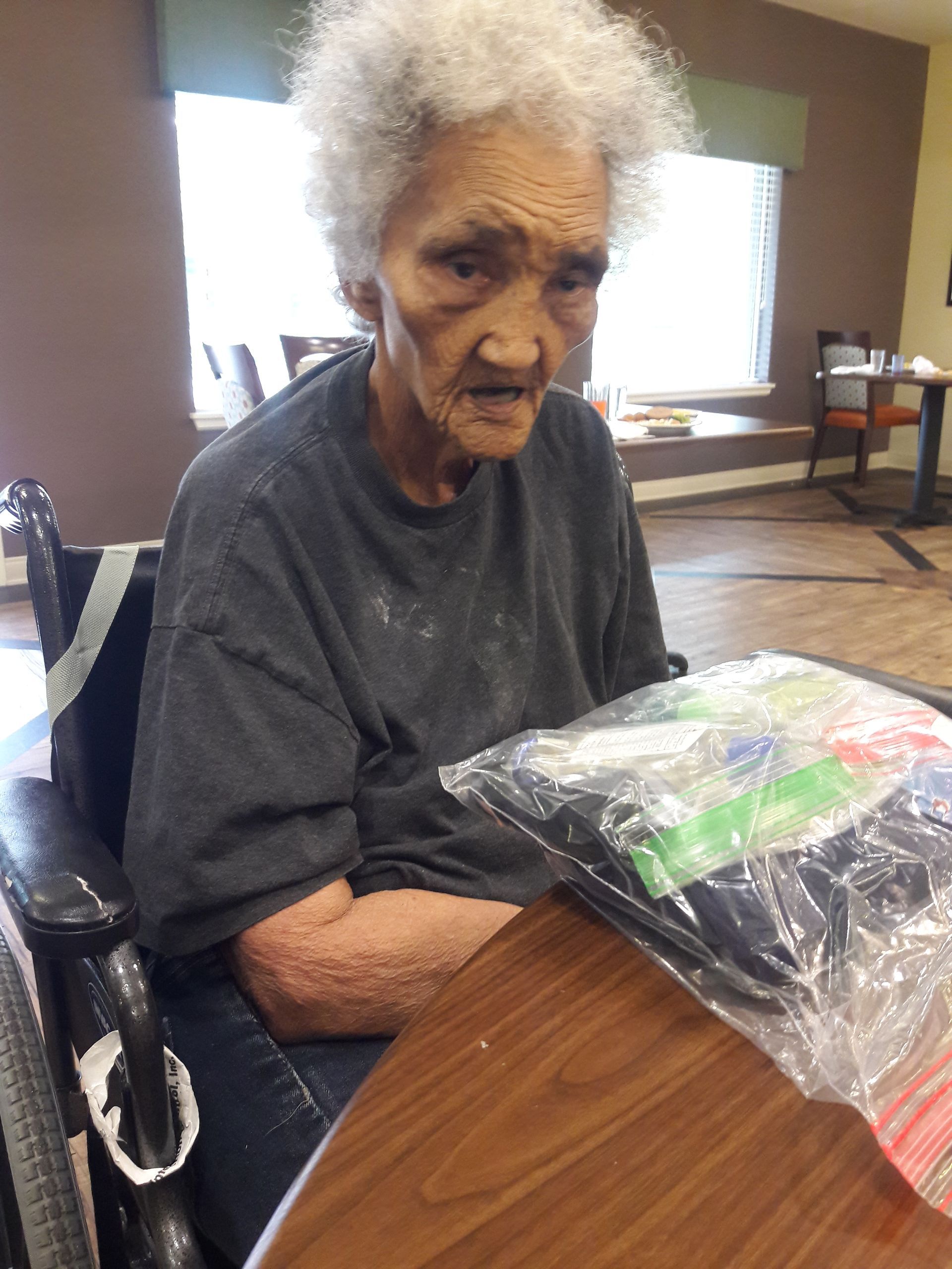 An elderly person in a wheelchair at a table, holding a plastic bag. They appear to be in a facility or common area.