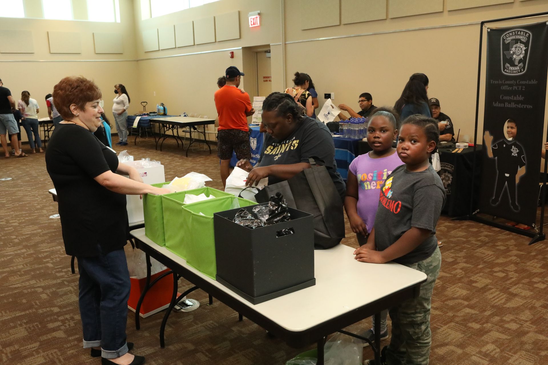 People at a community event, one woman serves items from containers. Two children and other attendees are present.