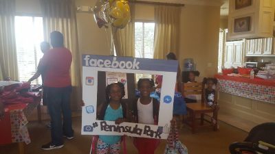 Two children posing inside a Facebook-themed photo frame at Peninsula Park, with adults and decorations in the background.