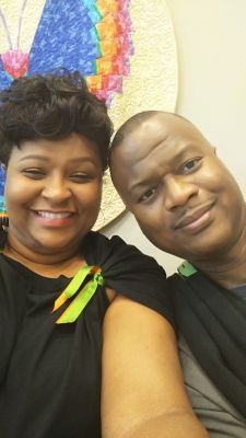 Smiling people pose for selfie, woman in black dress, man with shaved head, colorful background.