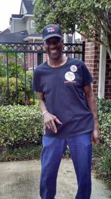 Person wearing a black hat and shirt with a button, standing outside by a fence and bushes.
