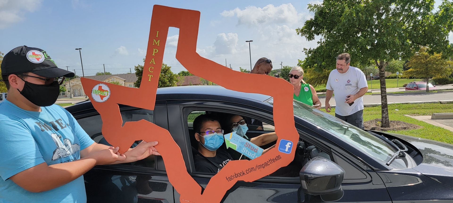 People holding a large Texas-shaped sign, handing something to people in a car. All are wearing masks.