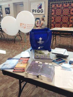 Table display: Brochures, balloons, and tote bag advertising an event in a room with a large quilt.