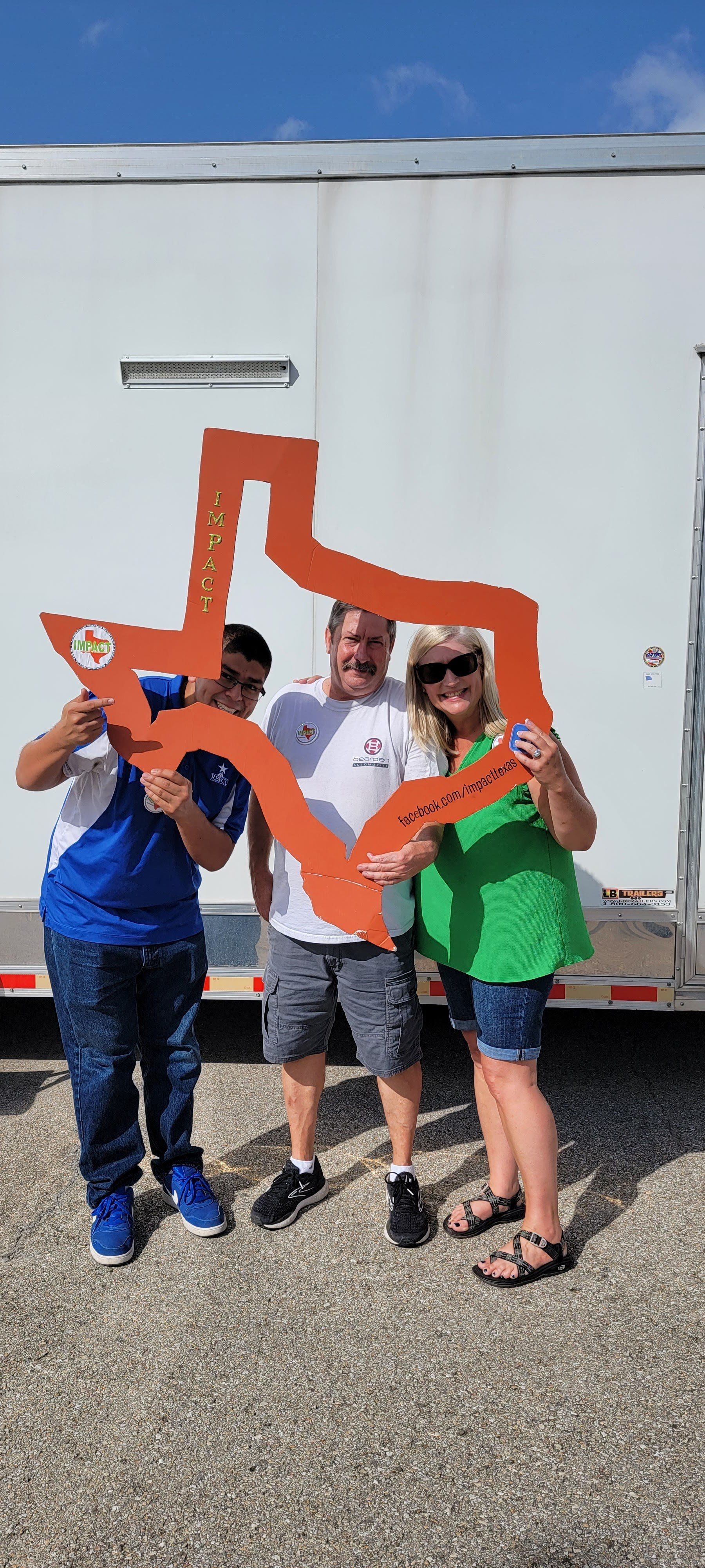 Three people pose with a Texas-shaped cutout. They stand in front of a white trailer on a gravel surface under a blue sky.