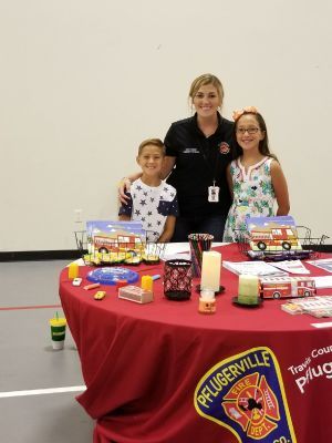 A fire department employee with two children at a table with fire safety themed items and Pflugerville logo.