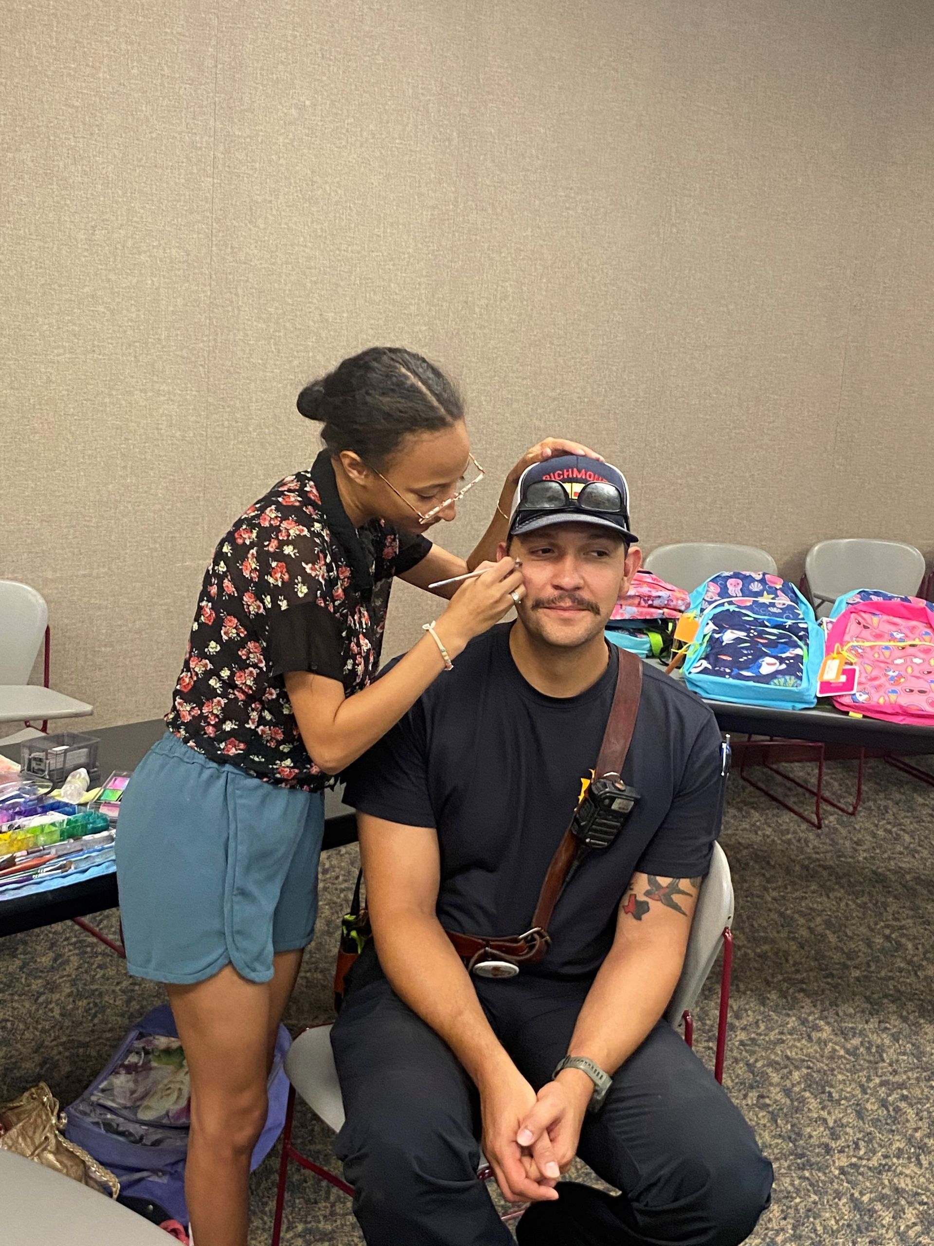 Woman adjusting a firefighter's hat while he sits, wearing a dark blue shirt and backpack harness; indoor event.