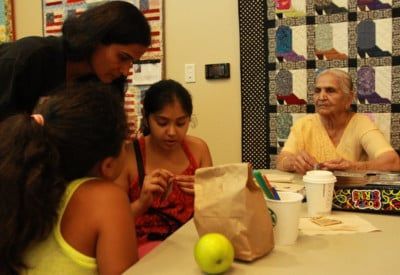 Group of people gathered at a table; woman in yellow sits, others lean in. Craft supplies are visible.
