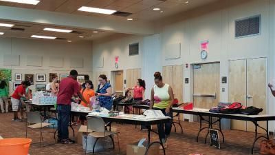 People at tables inside a room, possibly at a community event. Tables hold supplies; individuals interact.
