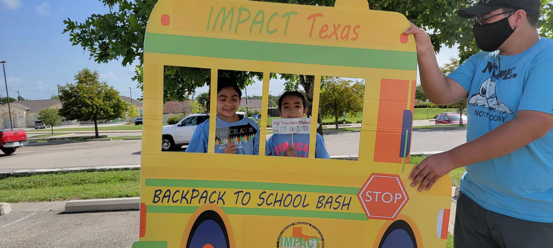 Two children posing in a school bus cutout, held by a person. Outside on a sunny day.
