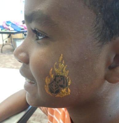 A child smiles, with a face tattoo of burning dice in orange and black.