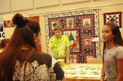 Two people view a vendor's table with a Christmas-themed quilt backdrop. T-shirts are on display.