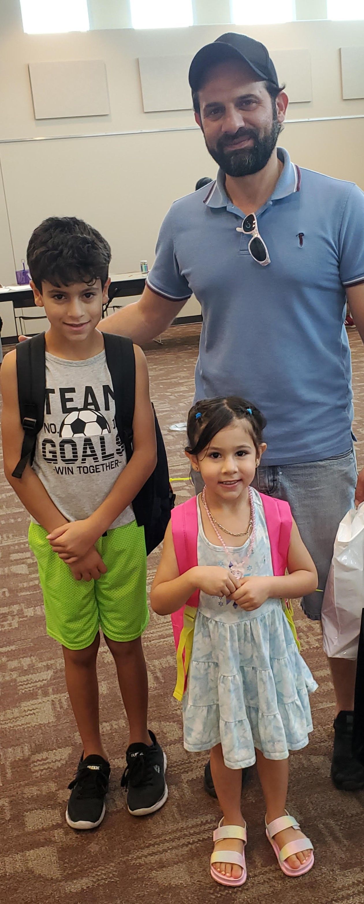 A man and two children pose indoors. The children wear backpacks, one in green shorts.