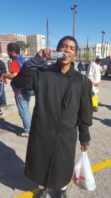Person drinking from water bottle, wearing a long black coat and holding a bag in a parking lot.