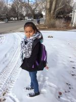 Girl stands in the snow, wearing a scarf, jacket, and backpack, on a snowy path.