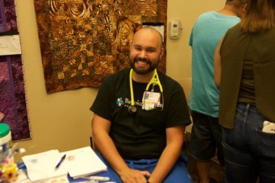 Man with stethoscope, smiling, at a medical station, wearing a black shirt, and surrounded by people.