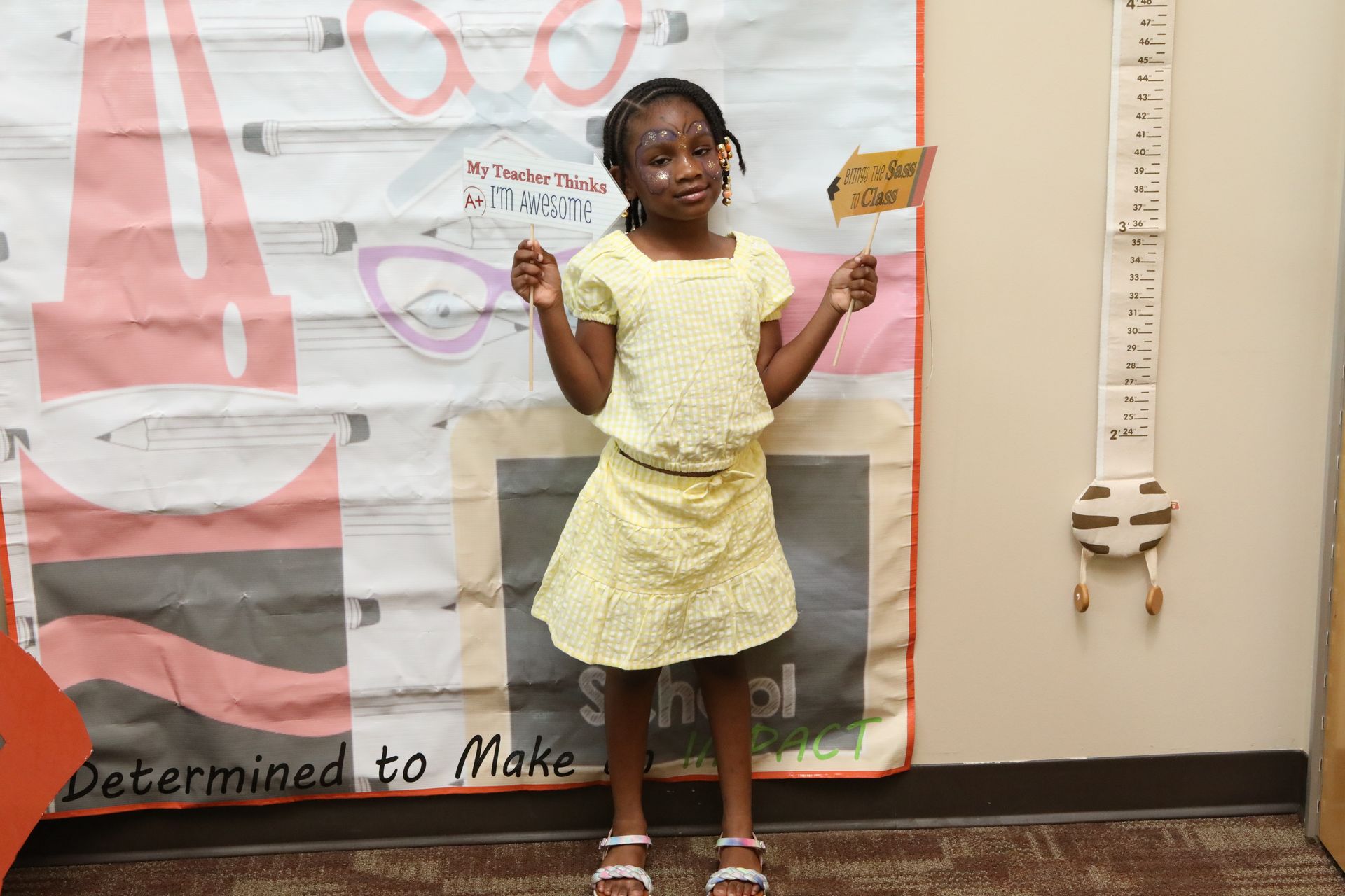 Girl in yellow dress, holding sign, stands in front of a height chart and wall decorations.