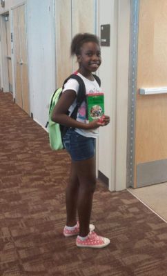Girl with backpack holding a book, smiling. In a hallway with wood doors and brown carpet.