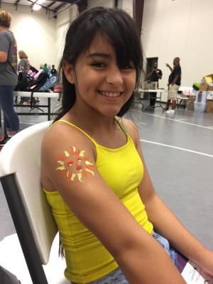 Girl with a painted sun on her arm smiles in an event hall, wearing a yellow tank top.
