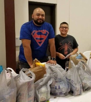 Man in Superman shirt and boy smile beside food bags on a table.