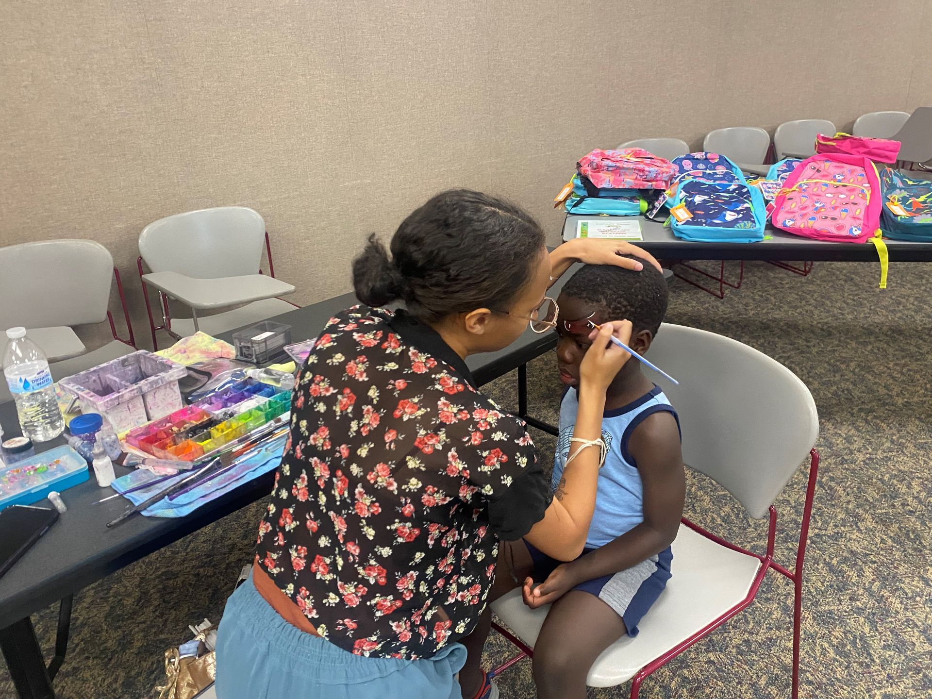 Person painting a design on a child's face. Child sits in chair, looking down. Backpacks are on a nearby table.