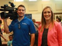 A cameraman with a camera and a woman smiling in front of a busy event.