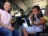 Two children inside a fire truck, one smiling, the other holding a green object, making a peace sign.