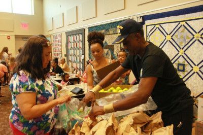 Man hands bags to woman at a community event. Other people are in the background, near a quilt display.
